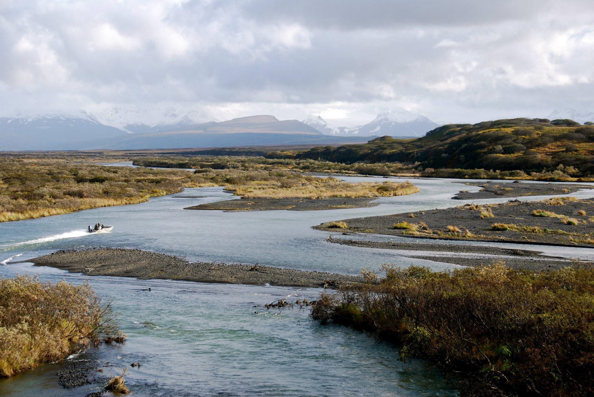 A boat speeds down a winding river surrounded by brush and mountains under a cloudy sky