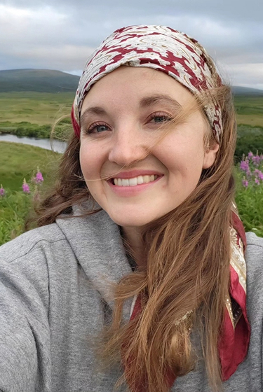 Smiling woman in gray hoodie and patterned headscarf with scenic river backdrop