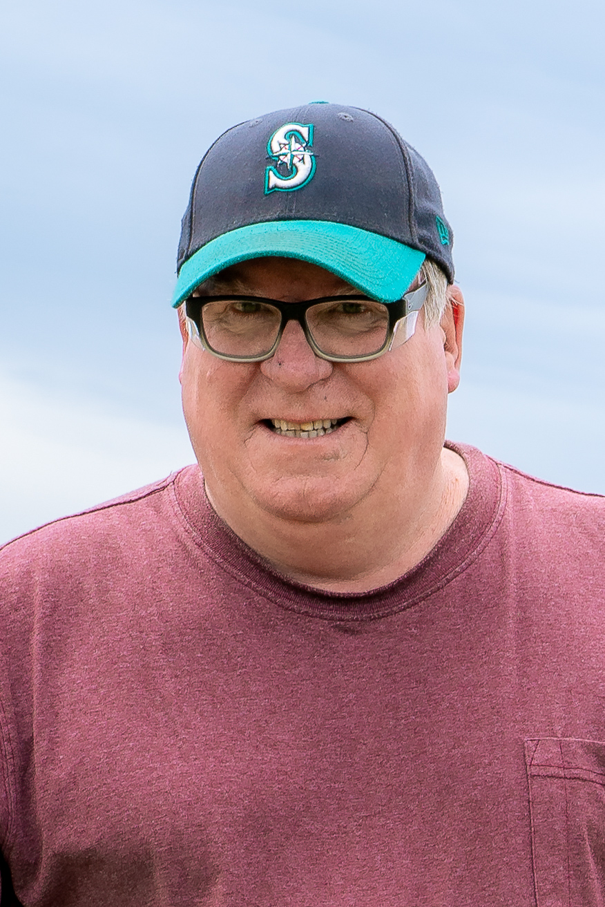Close-up portrait of smiling man wearing glasses, maroon shirt, and baseball cap at Alaska fishing camp