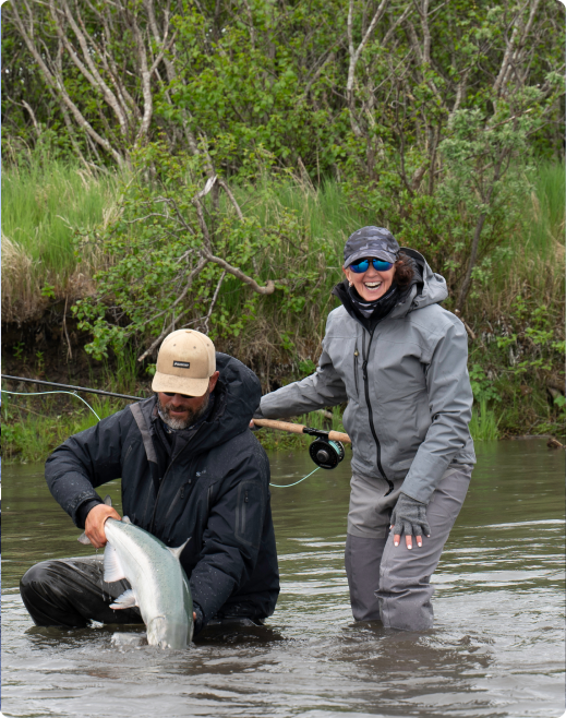 Two anglers in waders, one holding a large fish while the other smiles, standing together in a river