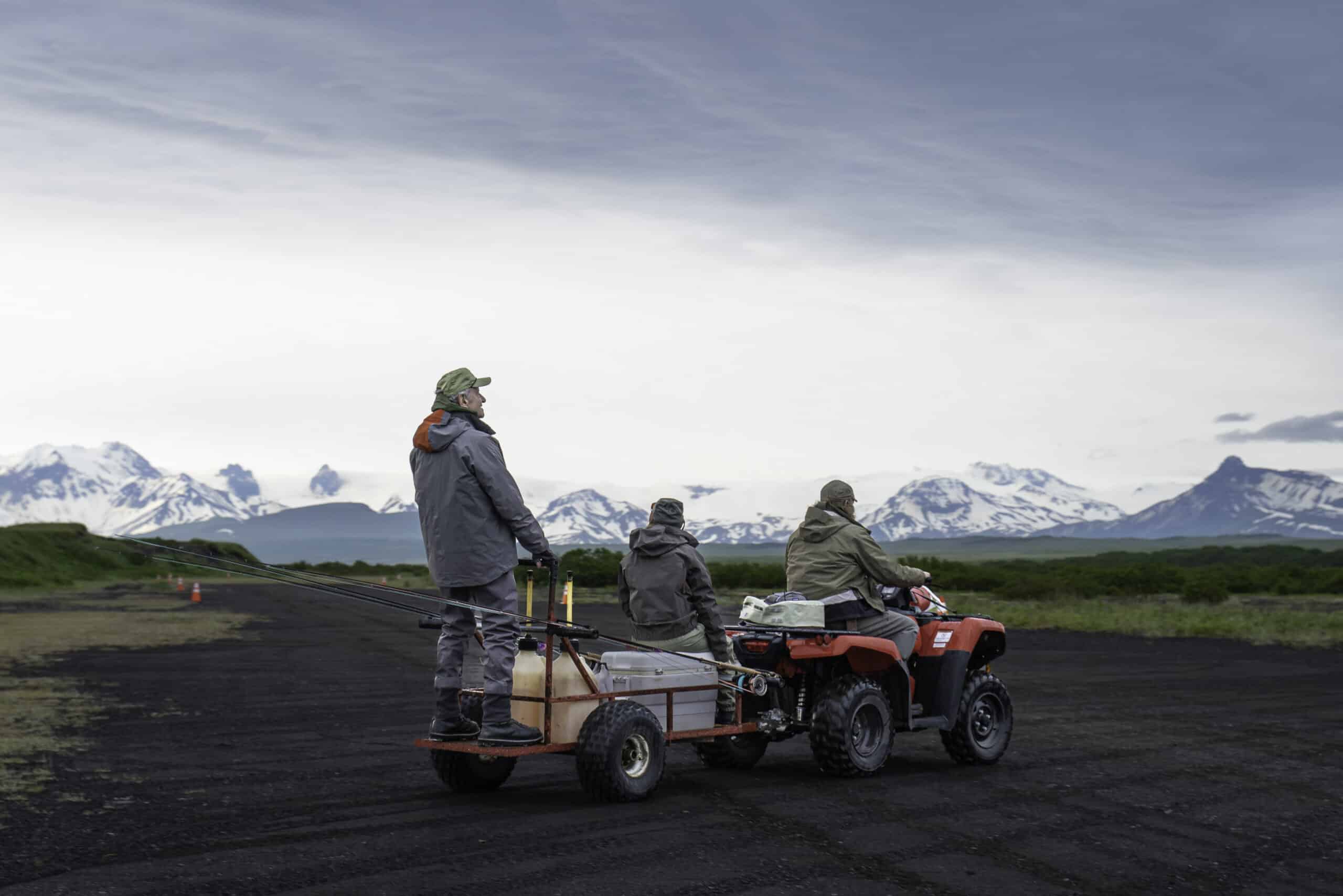 Anglers ride ATVs across Alaska tundra with fishing rods and snowy peaks in the background