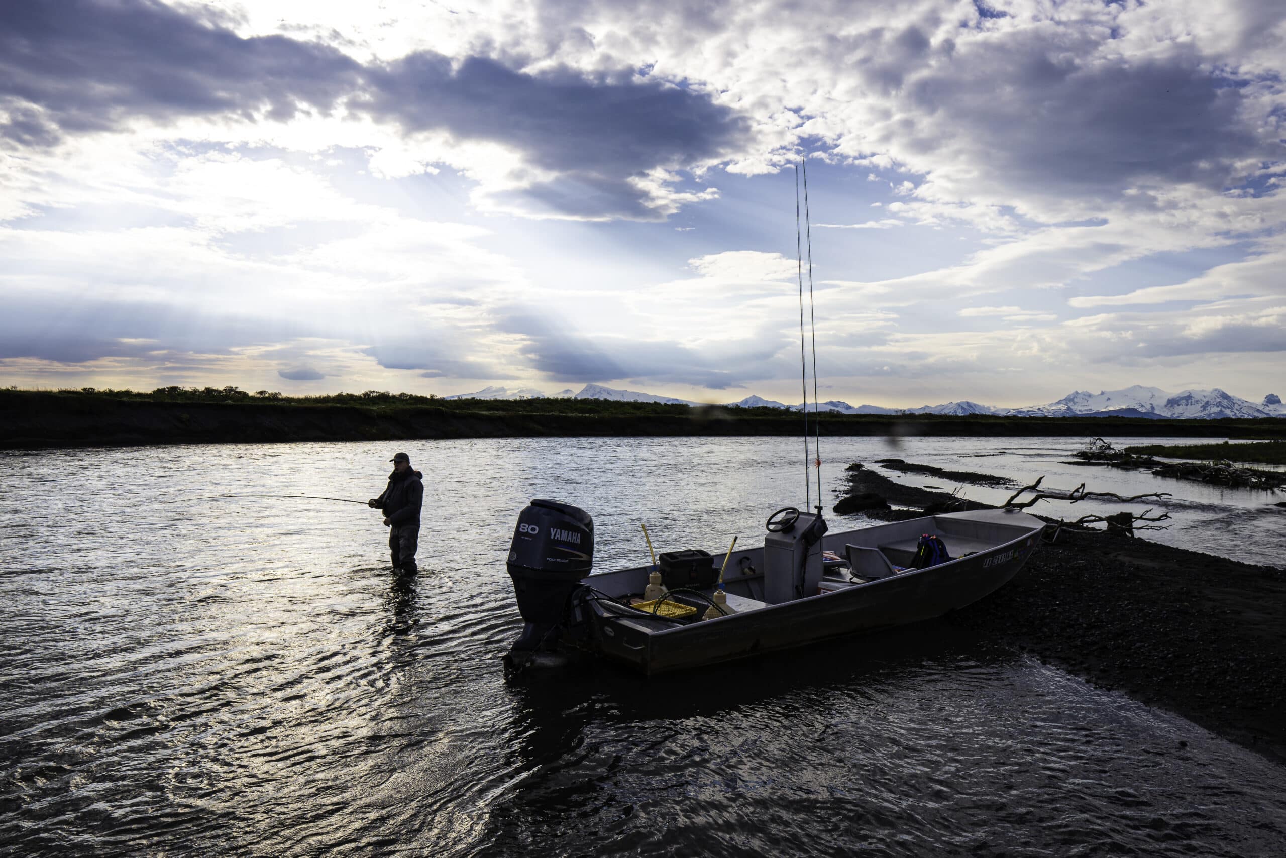 Angler fly fishing at sunset beside a riverboat with Alaska mountains in the distance