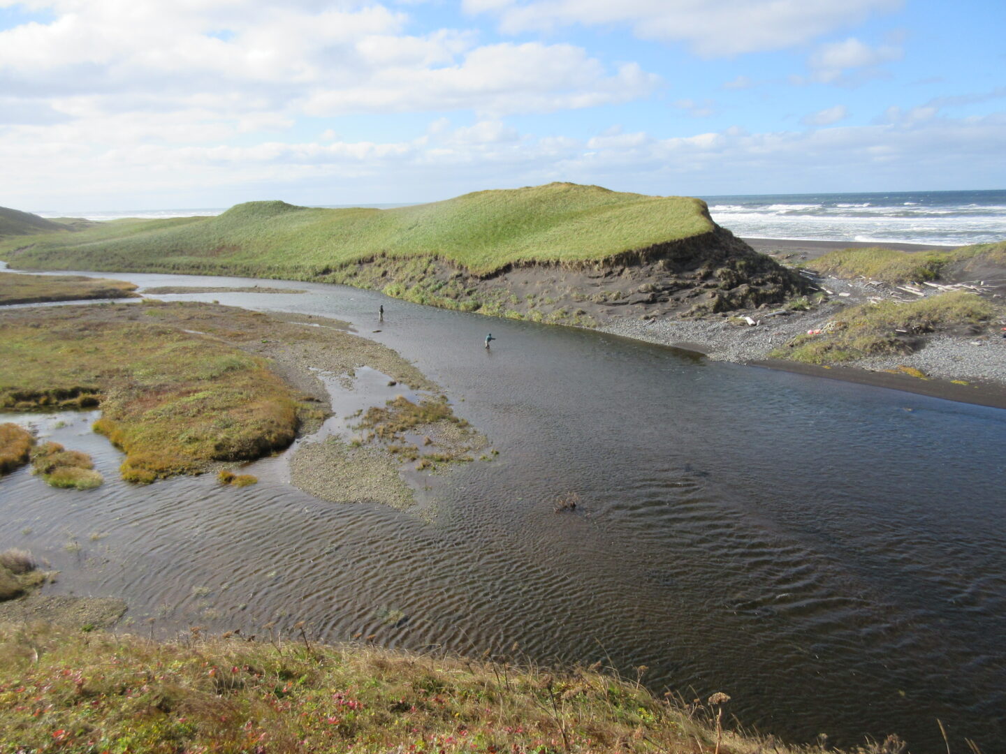 Two anglers wade in a winding river near grassy bluffs, with ocean waves visible in the background