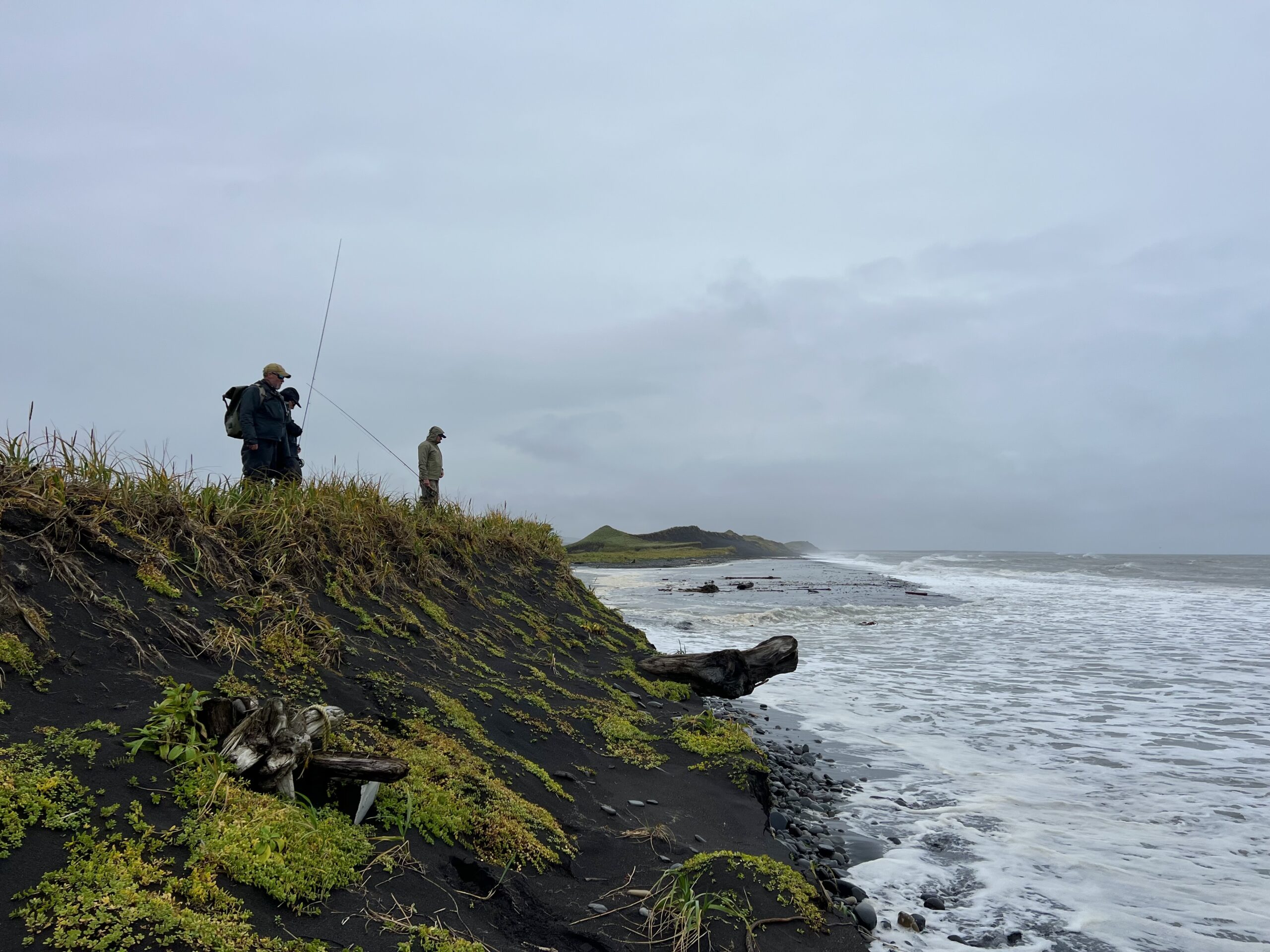 Two anglers stand on a grassy bluff above the ocean, casting lines toward waves under a cloudy sky