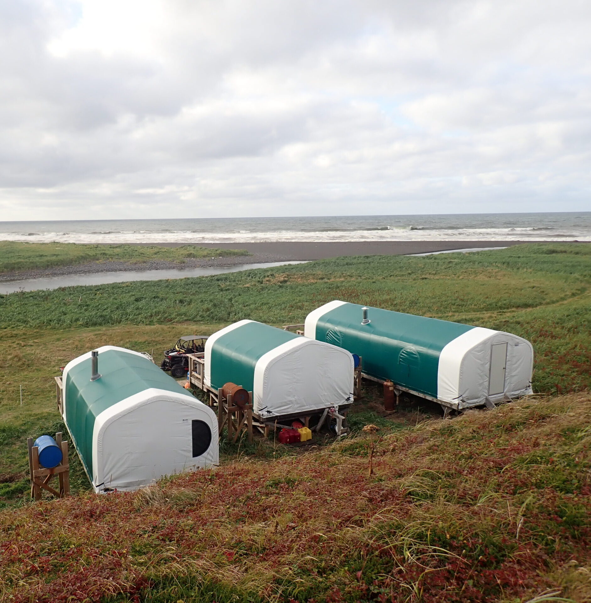 Three green-and-white camp tents overlook the ocean, set on grassy land near the beach