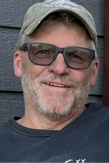 Man with a beard wearing glasses and a cap, smiling while sitting against a dark wooden wall
