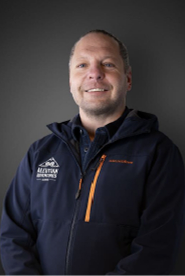 Man smiling in a navy jacket with orange zipper, logo on chest, standing against a dark background