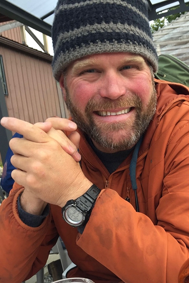 Smiling man in orange jacket and striped beanie sitting outdoors