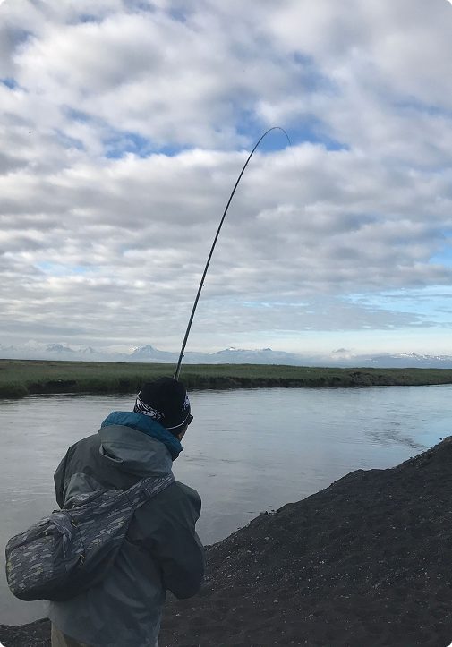 Angler with a backpack casts into a calm river under a cloudy sky, rod bent as they fight a fish