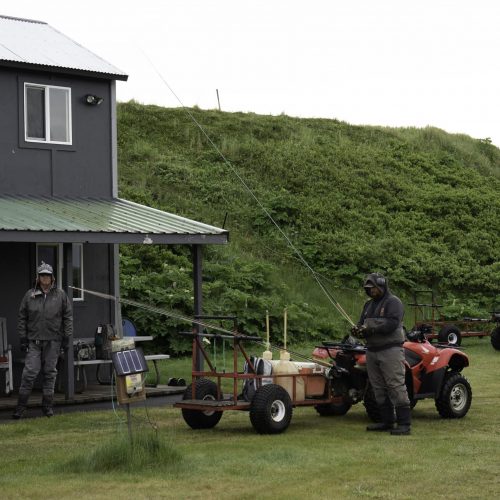 Anglers prepare gear by cabin with ATVs and fishing rods at a remote Alaska river camp
