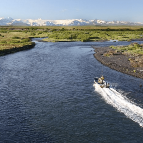 Boat traveling down a winding river with green fields and snow-capped mountains in the background