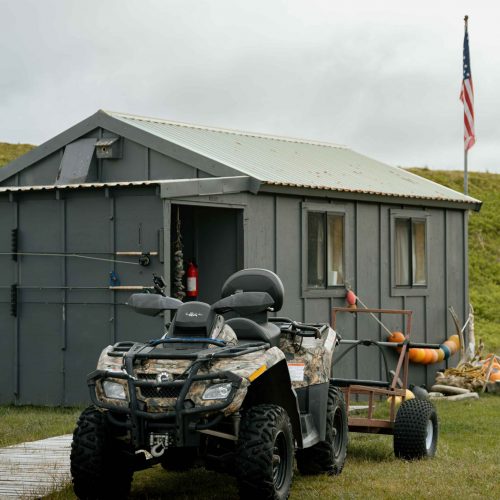 Small cabin with fishing rods on wall and ATV parked outside at Alaska river camp