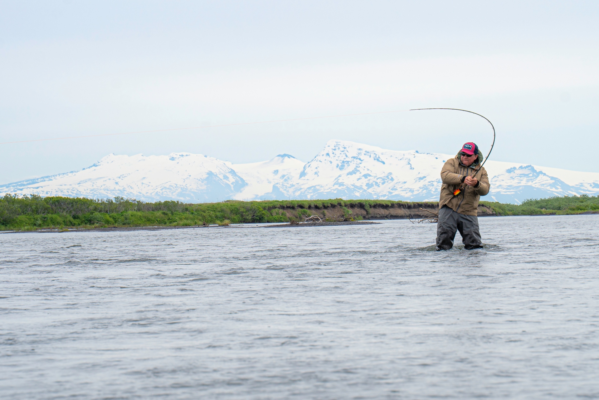 Angler fly fishing in Alaska river with snow-covered mountains in background
