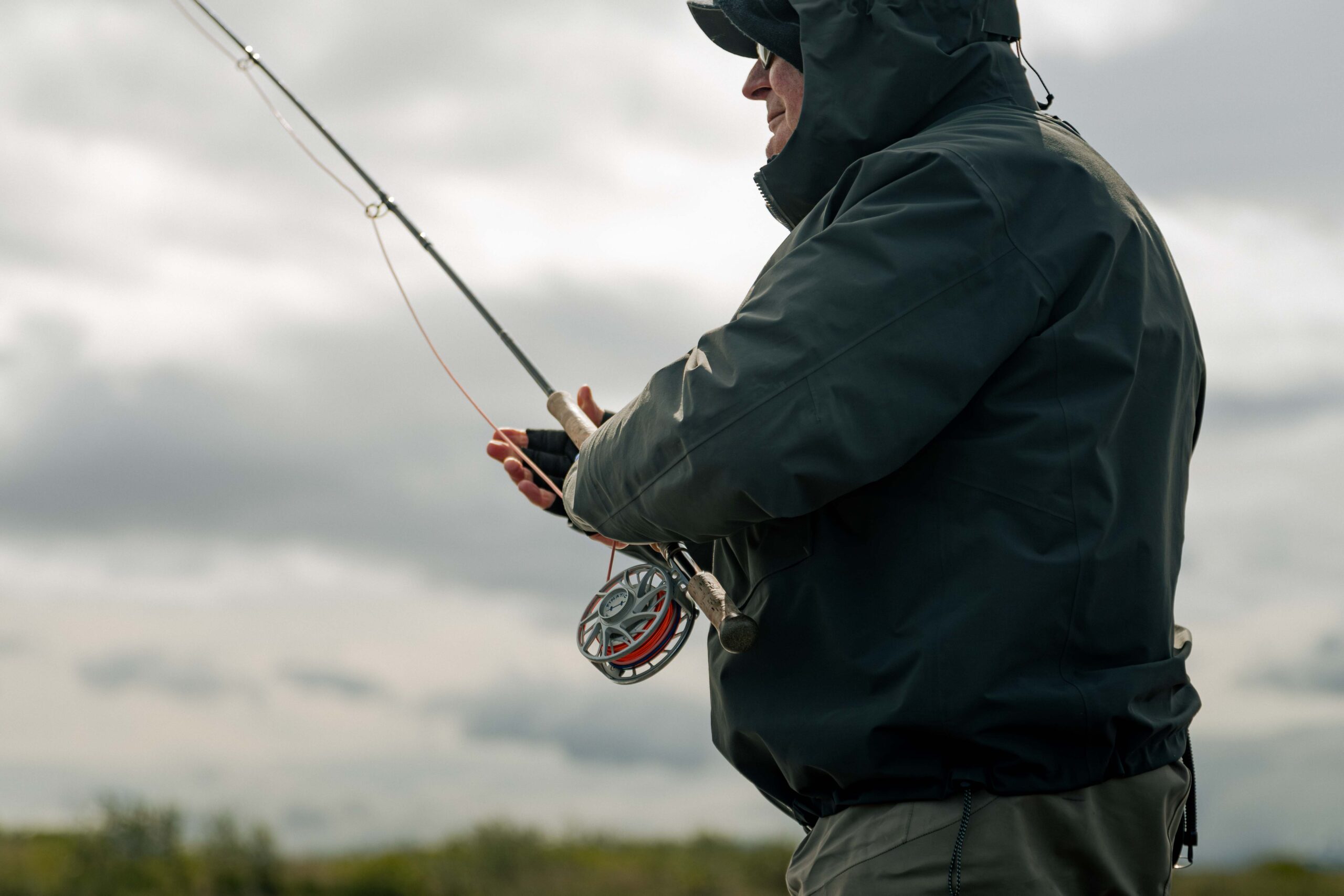 Angler in dark jacket holding fly fishing rod with reel against cloudy sky