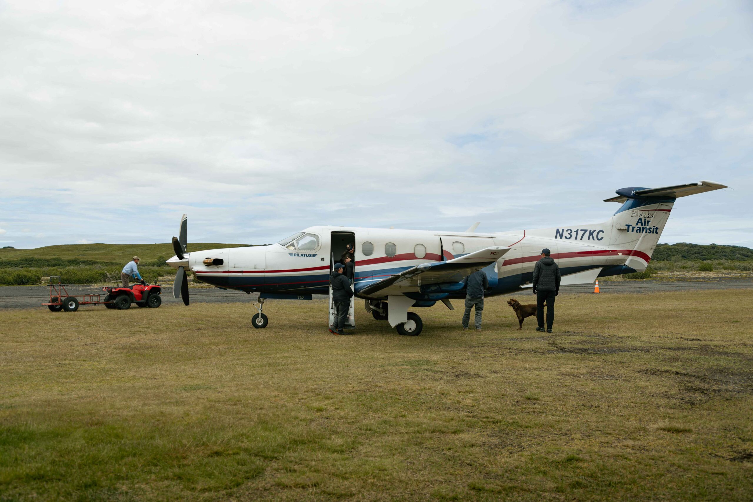 Small charter plane on rural Alaska airstrip with passengers, dog, and ATV nearby
