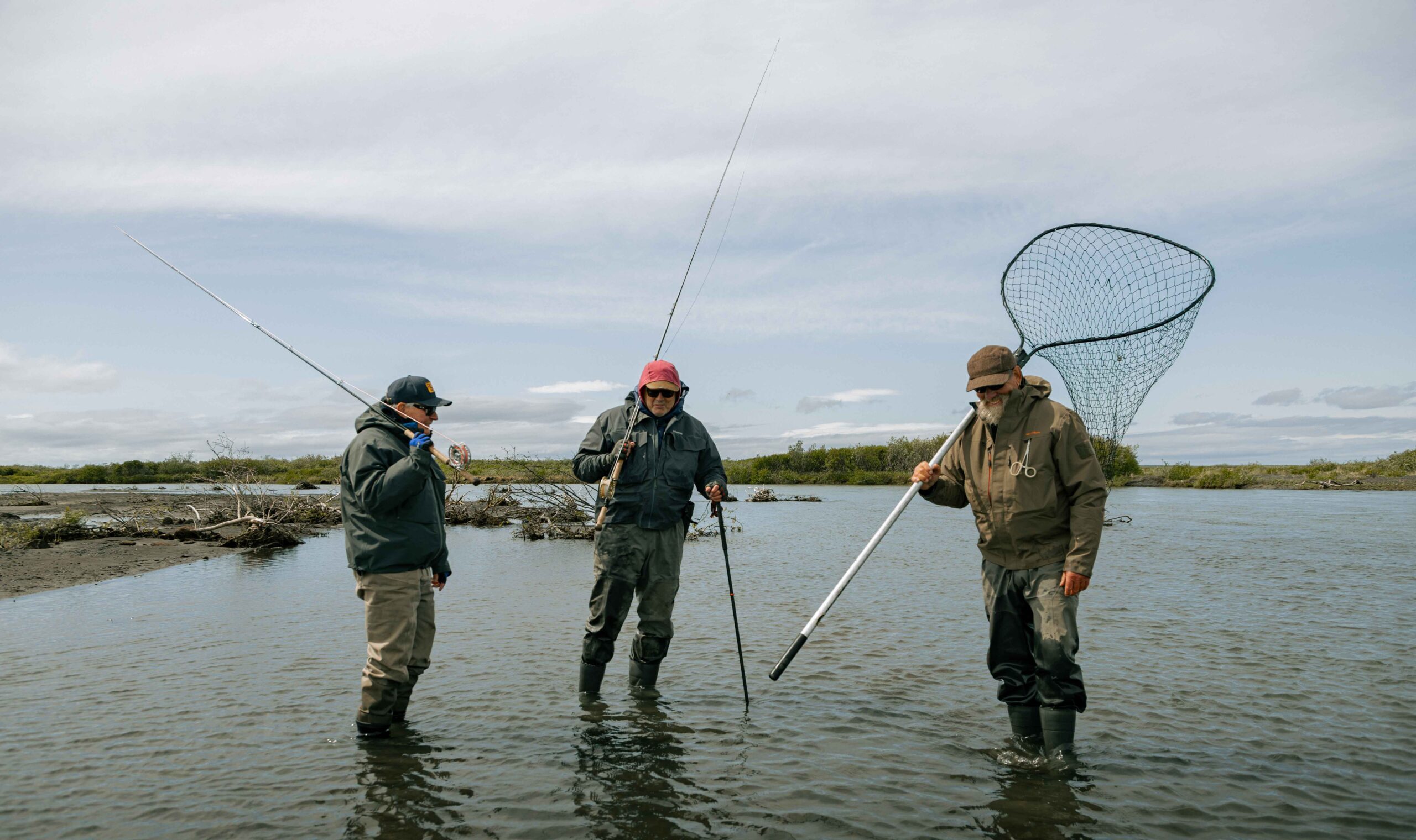 Three anglers standing in river shallows with fishing rods and large landing net
