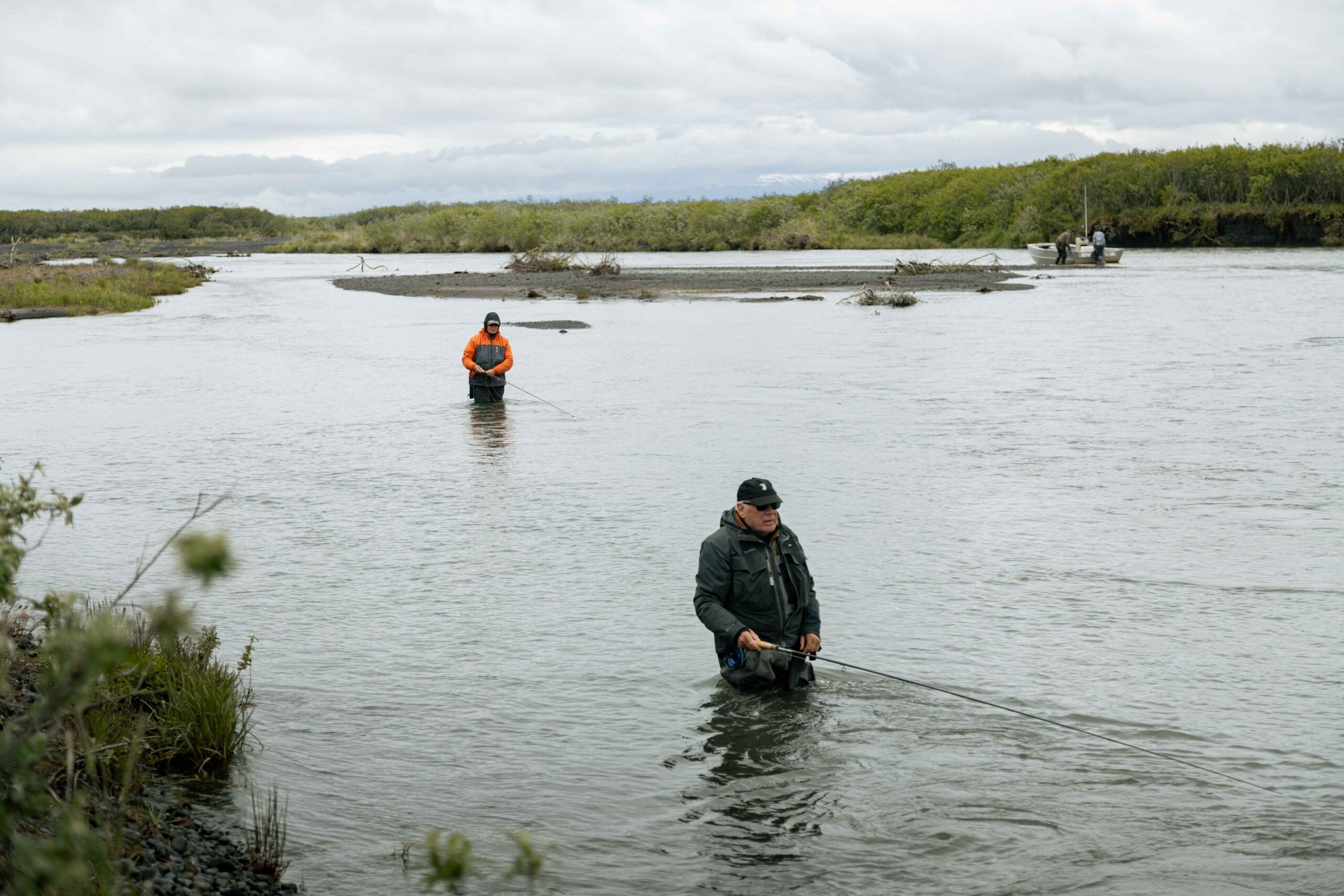 Anglers wading in wide river while fishing with rods, boat anchored in background
