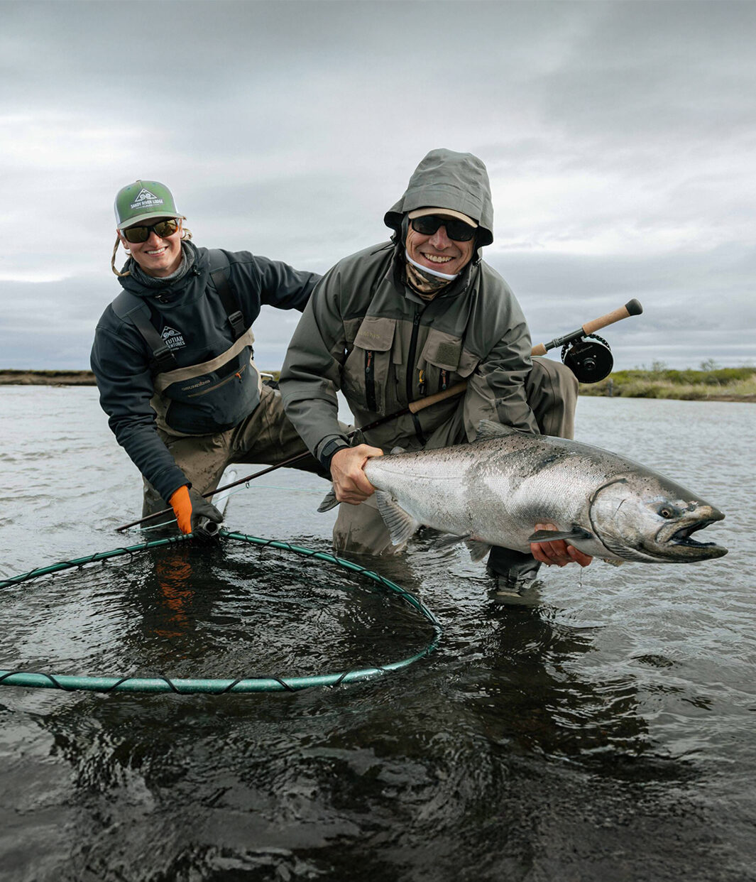 Angler holding large king salmon while another fisherman assists with landing net in river