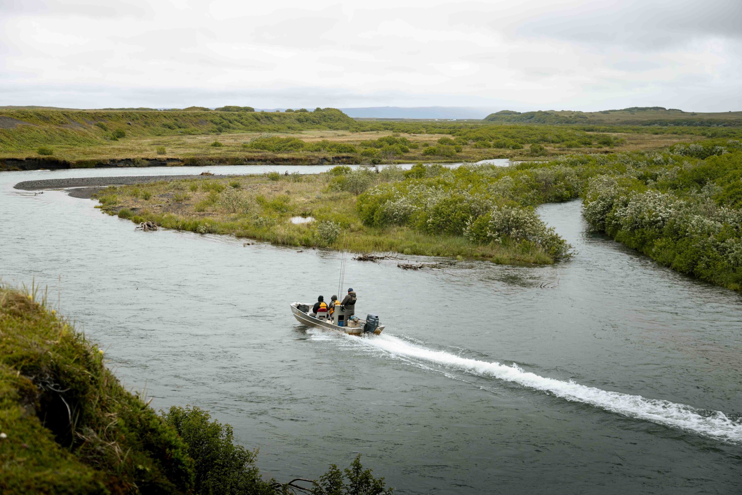 Small boat with anglers traveling down winding Alaska river through green tundra