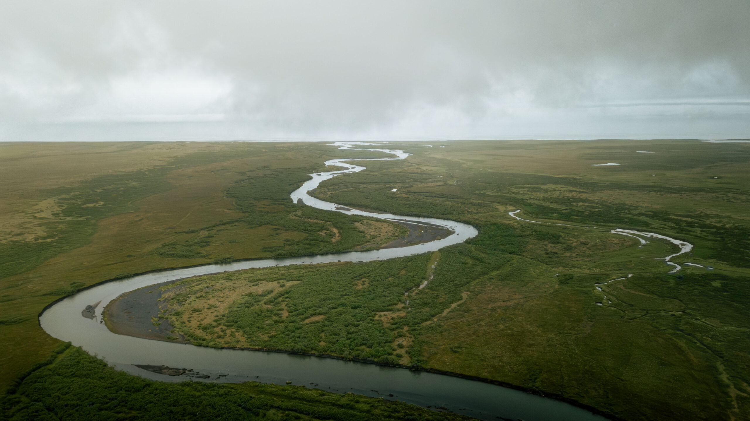 Aerial view of winding Alaska river cutting through vast tundra under cloudy sky